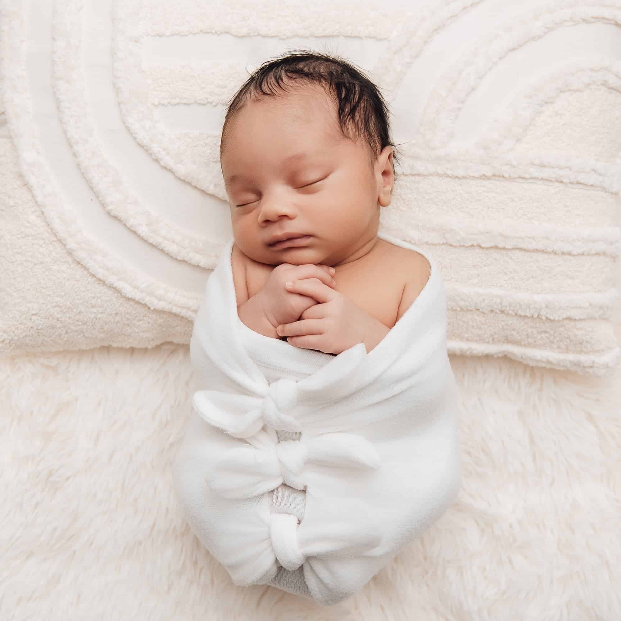 Séance photo bébé idées et préparation: jeune bébé endormi, enveloppé dans une couverture blanche nouée, posé sur un fond doux et moelleux.