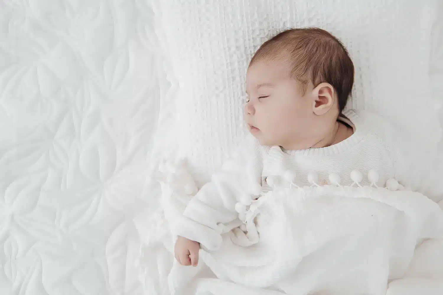 Bébé endormi sur lit blanc, vêtu d’un tricot blanc, dans un décor doux et lumineux.