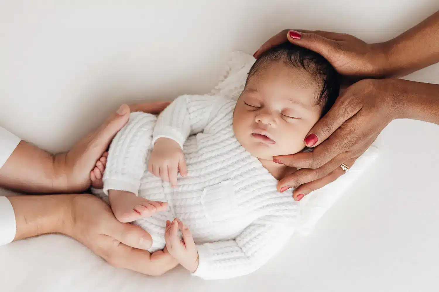 Bébé nouveau-né endormi dans une tenue blanche, entouré des mains des parents sur le ventre et la tête, dans une pose douillette.
