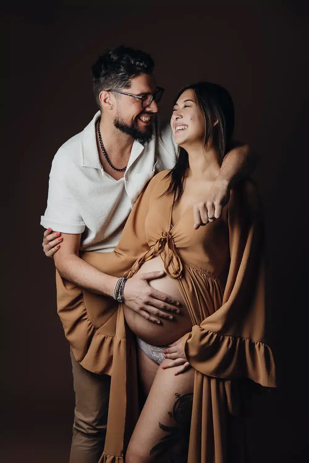 Couple heureux, la femme enceinte porte un long kimono beige et le homme la tient par la taille, sourire partagé, lumière douce sur fond sombre.