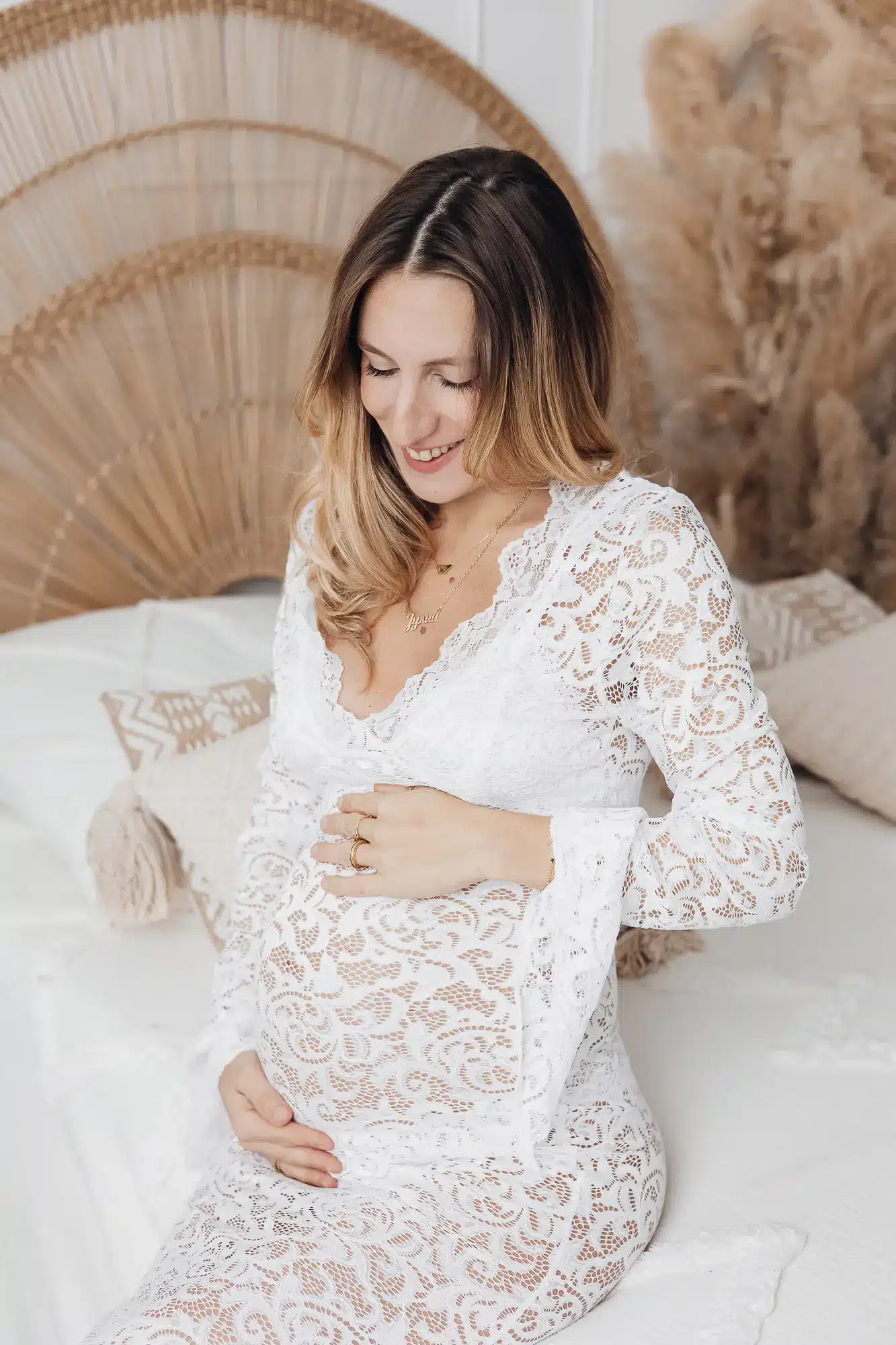 Femme enceinte assise sur un lit, vêtue d’une robe en dentelle blanche, mains sur le ventre, sourire doux dans un décor intérieur chaleureux.