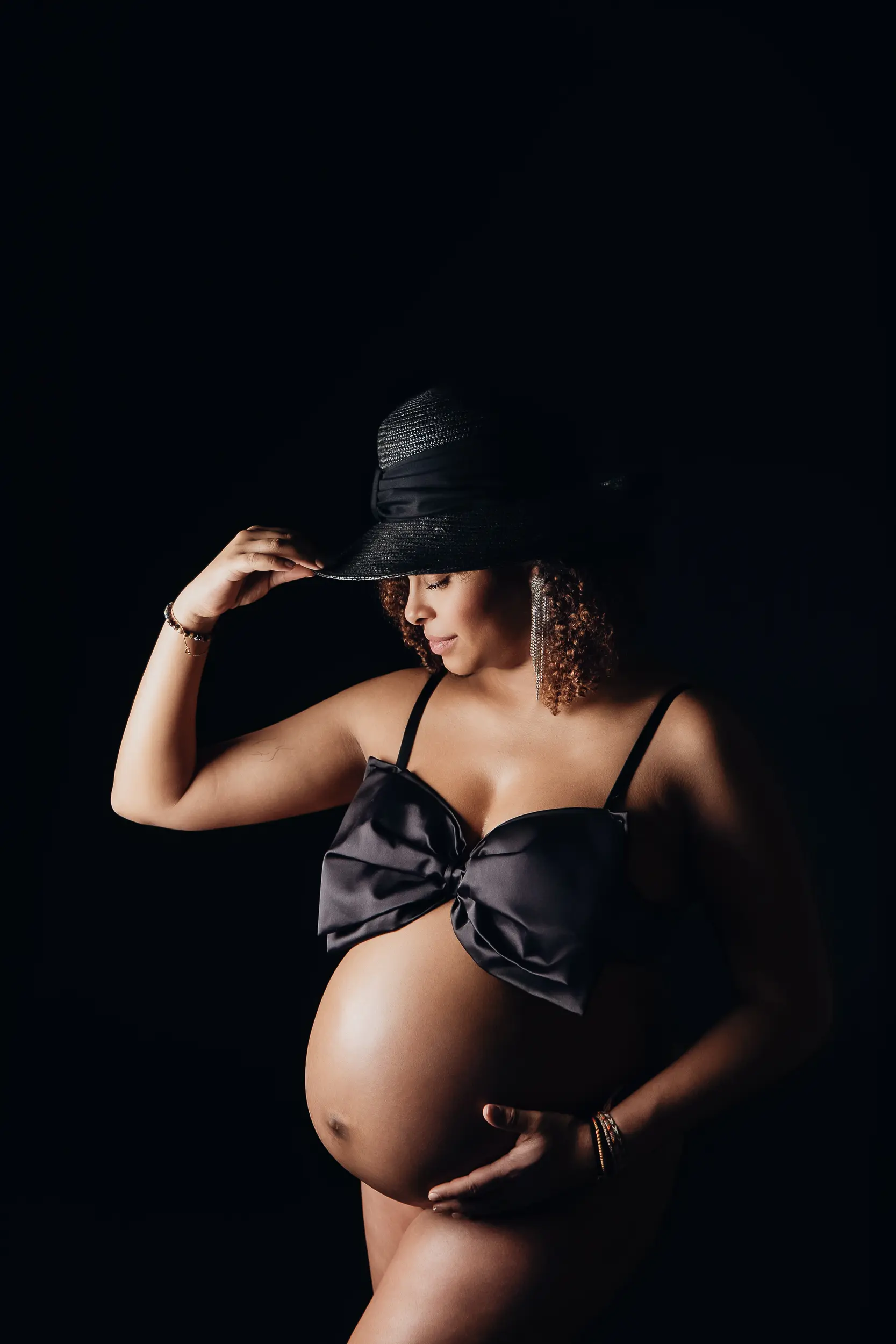 Femme enceinte portant un chapeau noir et un gros nœud sur le buste, mains sur le ventre rond, éclairage doux sur fond sombre.