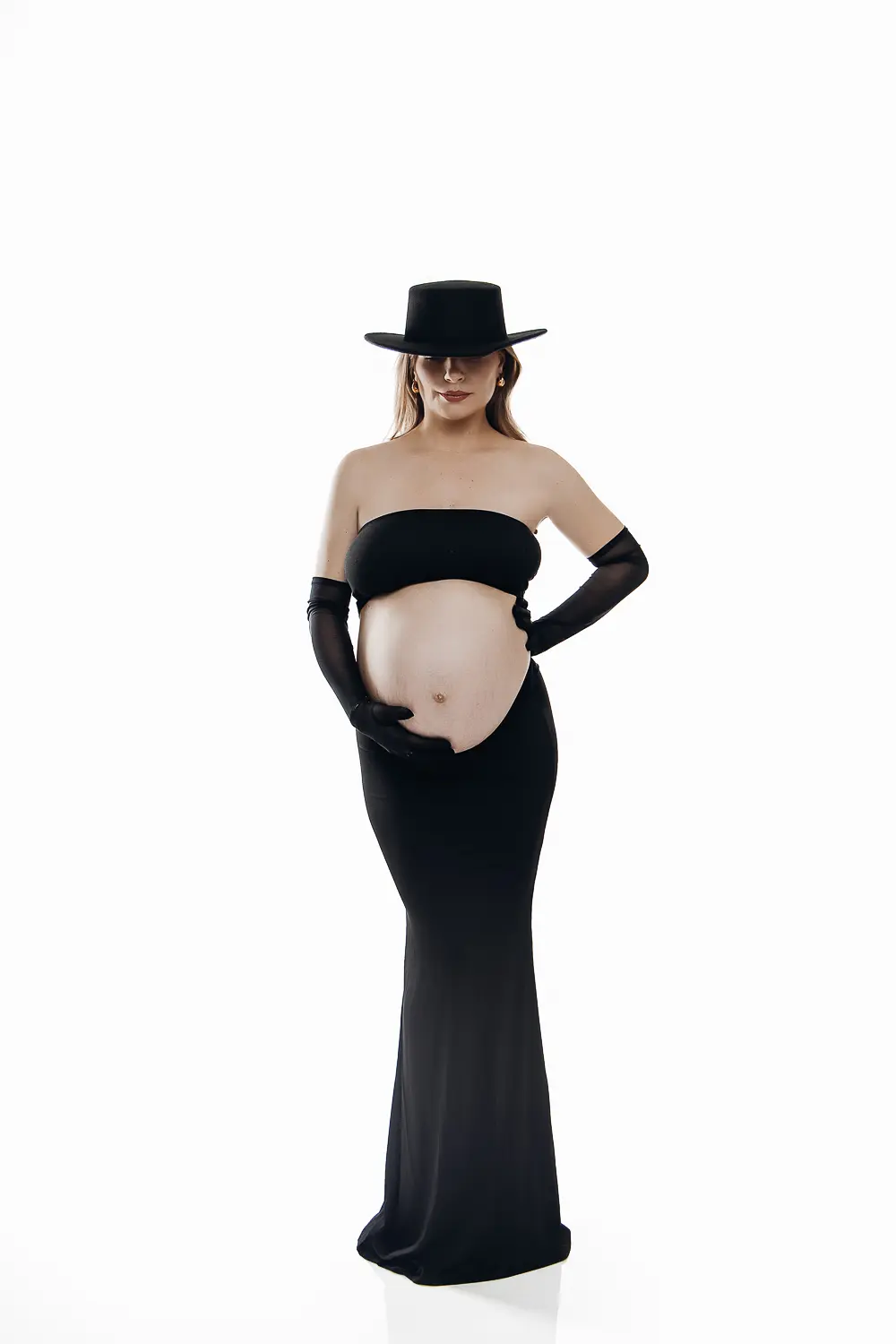 Femme enceinte en robe noire moulante et gants longs, portant un chapeau noir, posant les mains sur le ventre. ALT: maternité, grossesse.