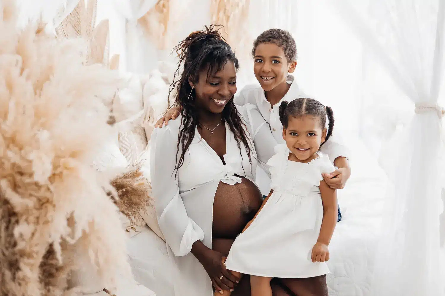 Femme enceinte souriante entourée de deux enfants dans un décor blanc et doux, pose mère-fille et enfant ado complices.