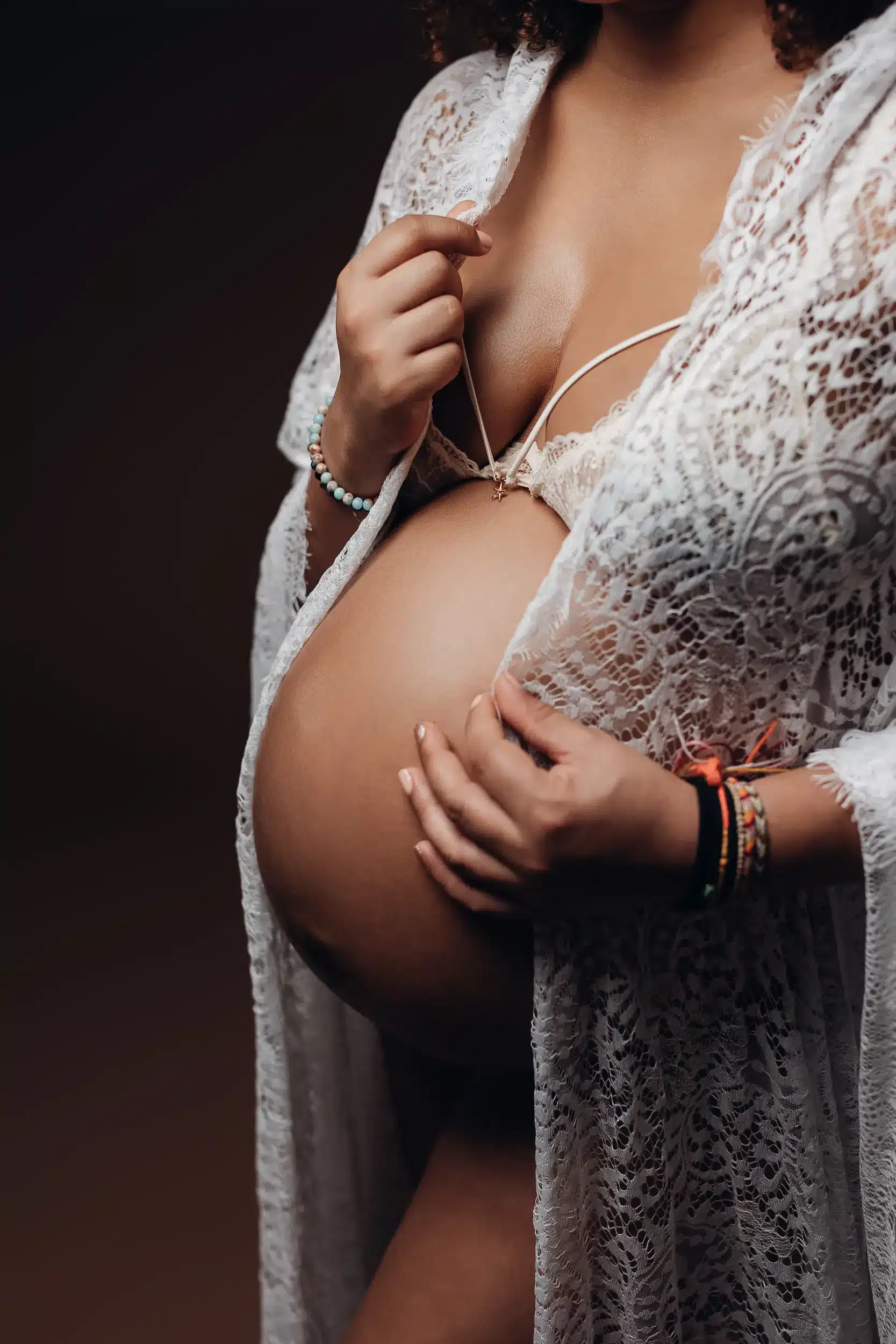 Gros plan sur une femme enceinte tenant son ventre rond, vêtue d’une robe en dentelle blanche, avec bracelets au poignet.