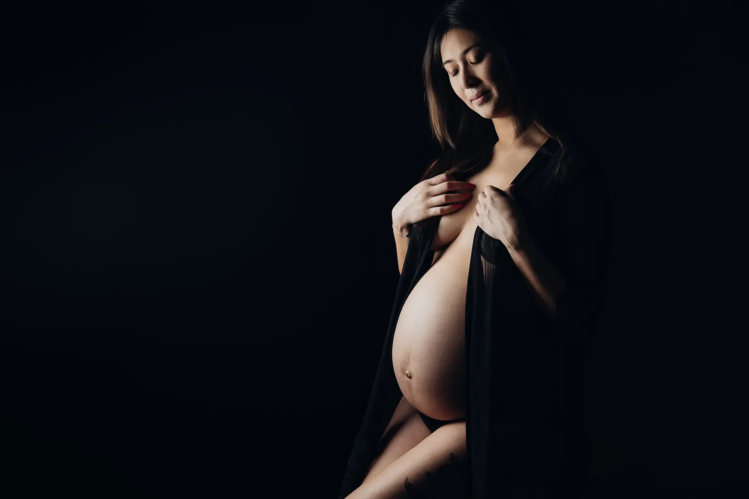 Portrait artistique d'une femme enceinte, seins nus partiellement couverts par un voile sombre, abdomen proéminent en lumière douce.