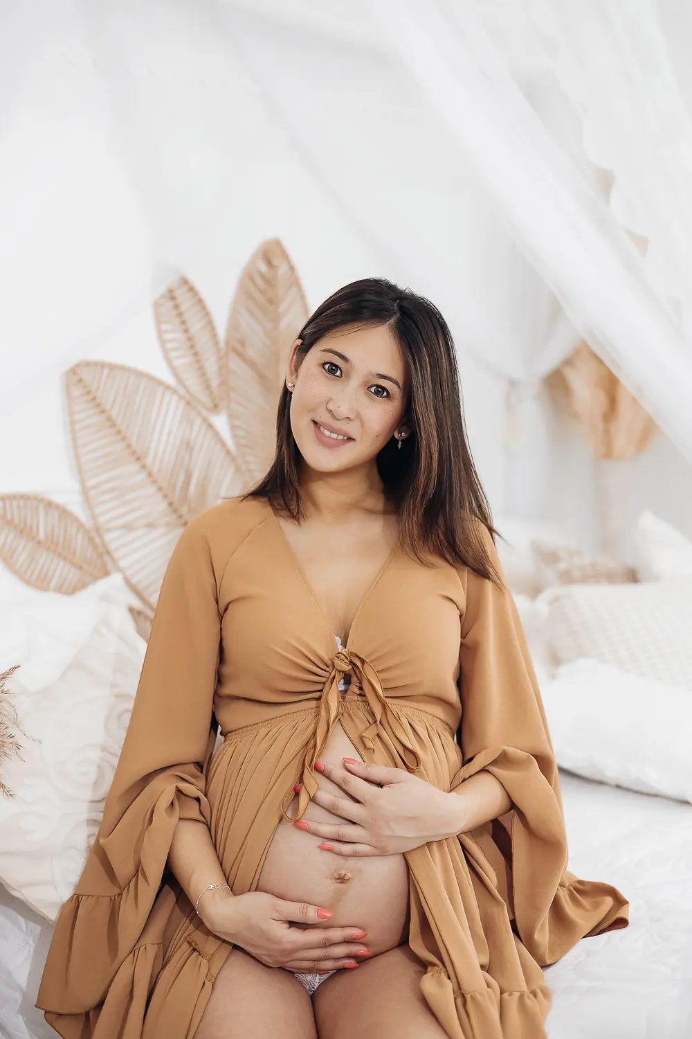 Portrait d'une femme enceinte souriante, ventre rond mis en valeur par une robe beige, mains posées dessus dans un cadre lumineux et doux.