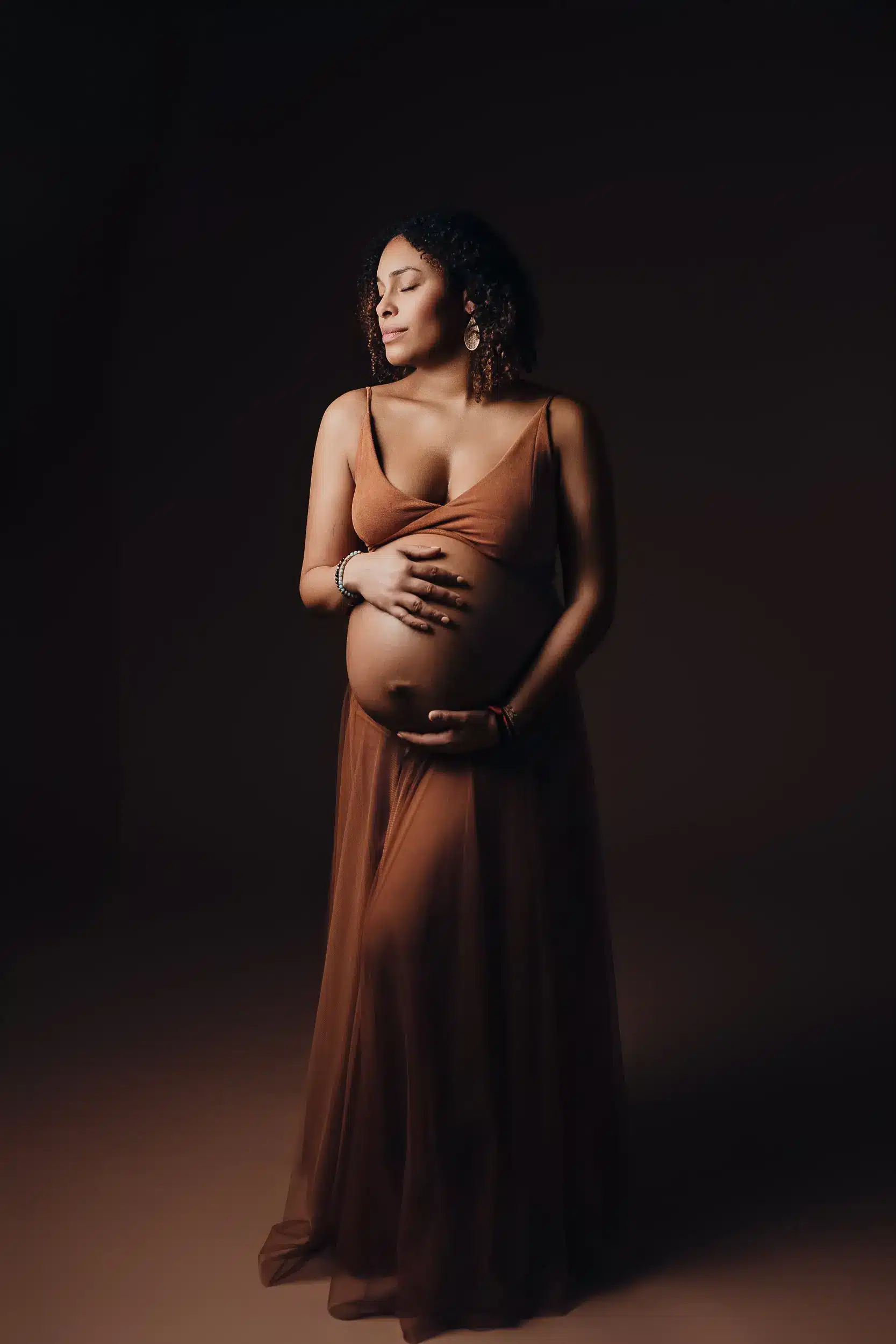 Portrait de femme enceinte en robe longue kaki, main sur ventre, éclairage doux sur fond sombre. Alt: femme enceinte posant lors d'une séance photo grossesse.