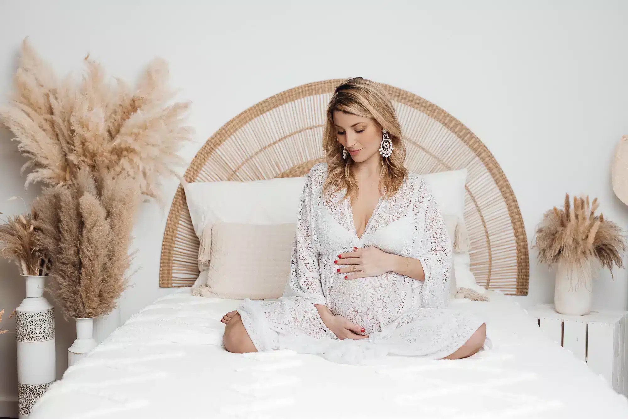 Alt text: Femme enceinte en robe blanche, assise sur un lit, main sur le ventre, dans une chambre lumineuse et décor bohème. pourquoi je ne suis pas photogenique ?