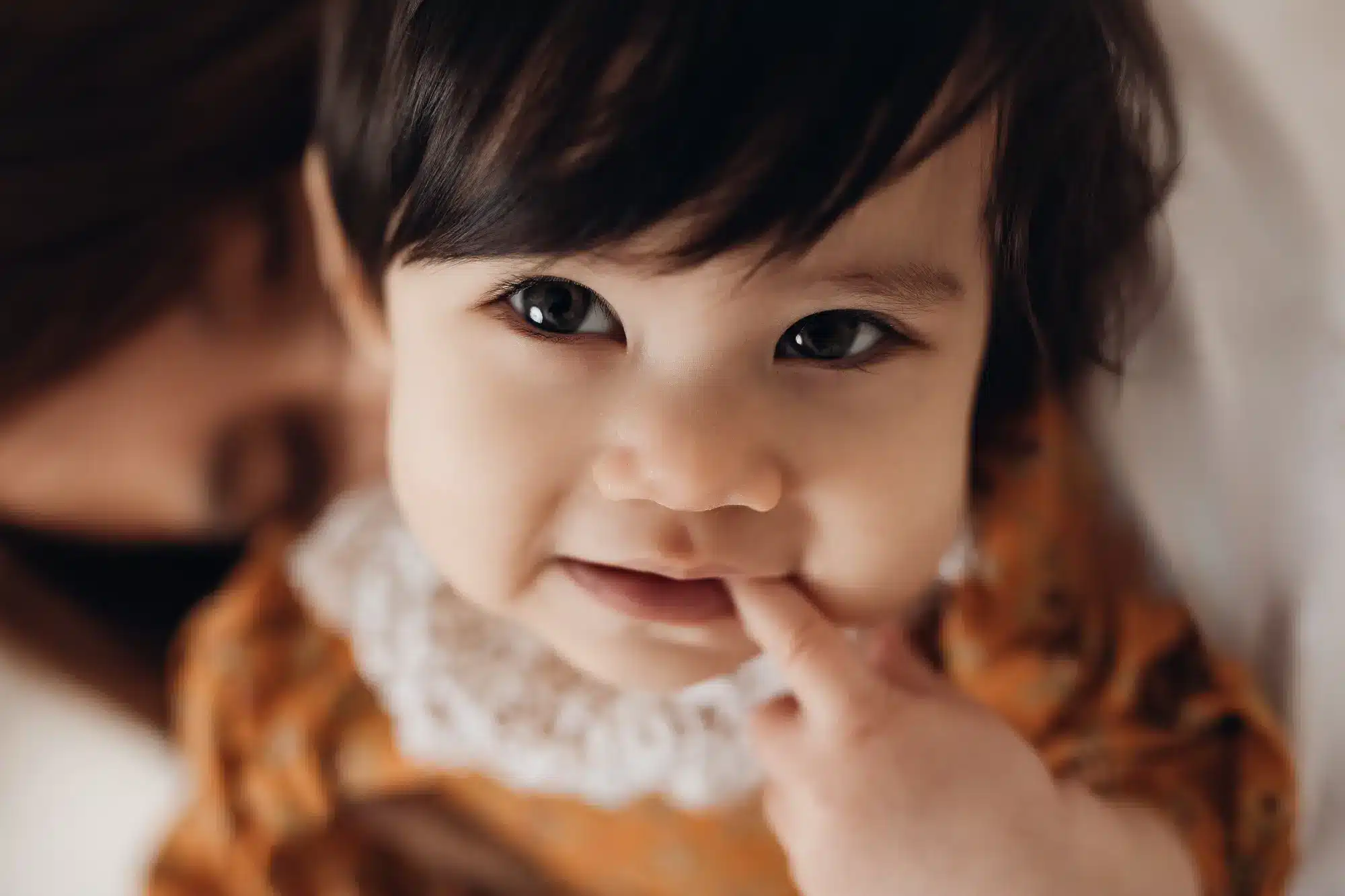 Bébé asiatique souriant, finger sur le nez, veste fantaisie orangée et collier blanc, portrait doux en lumière naturelle.
