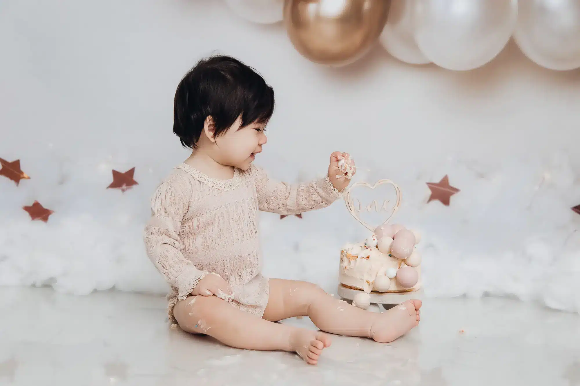 Bébé souriant assis sur le sol, tenue beige, jouant avec un gâteau et un décor pastel, ballon et étoiles au fond.
