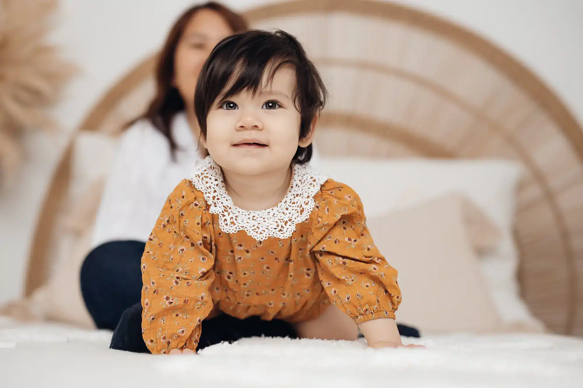 Enfant petit près d’un lit, assis sur une couverture blanche, vêtue d’une robe orange à imprimés, col en dentelle blanche, sourire doux.