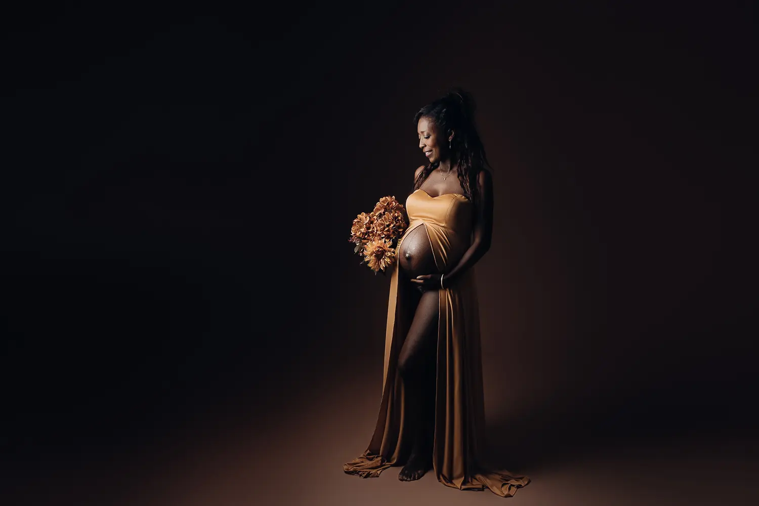 Future mère enceinte dans une robe dorée, pose portrait dans un studio sombre avec bouquet près du ventre.