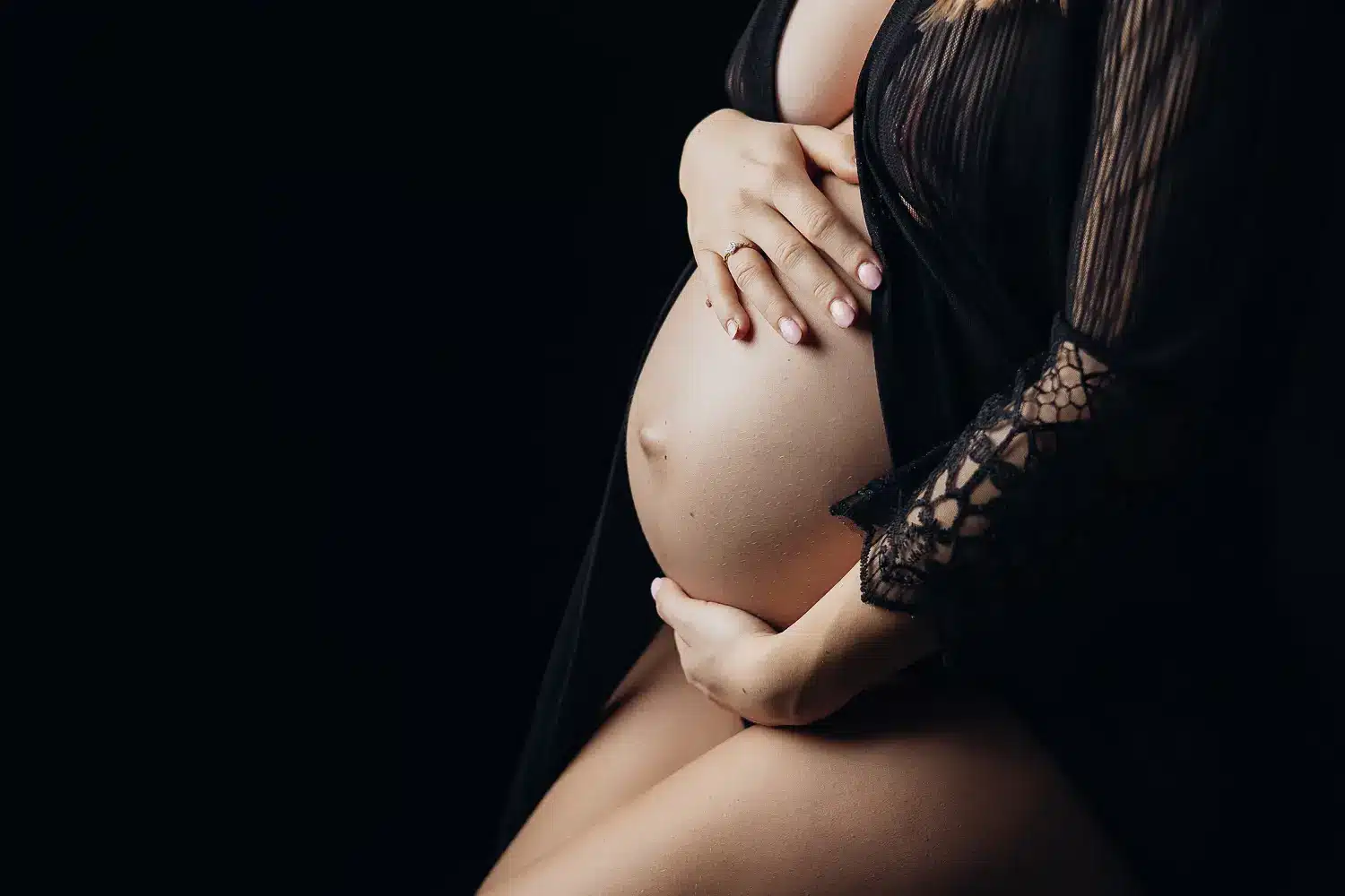 Gros plan sur une femme enceinte, ventre rond soutenu par ses mains, vêtement noir en dentelle sur fond sombre. ALT: Femme enceinte caressant son ventre, moment intime de grossesse.