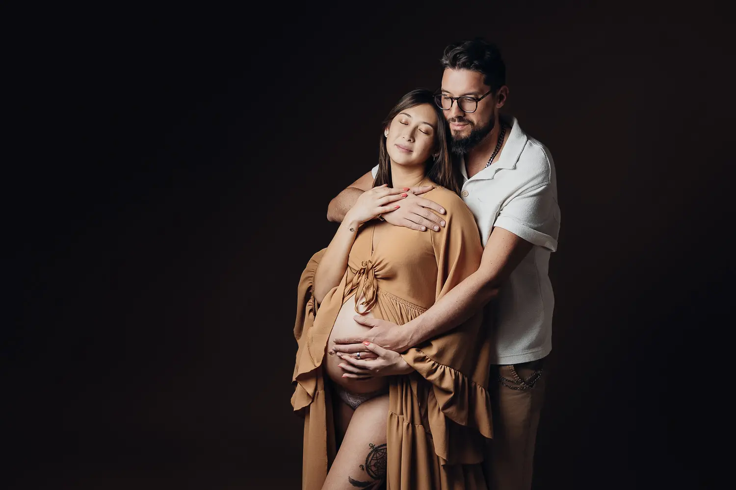 Grossesse: couple en studio, femme enceinte en poncho camel sur le ventre, homme en polo clair derrière elle, ambiance douce et lumineuse. Grossesse: couple en studio, femme enceinte en poncho camel sur le ventre, homme en polo clair derrière elle, ambiance douce et lumineuse.