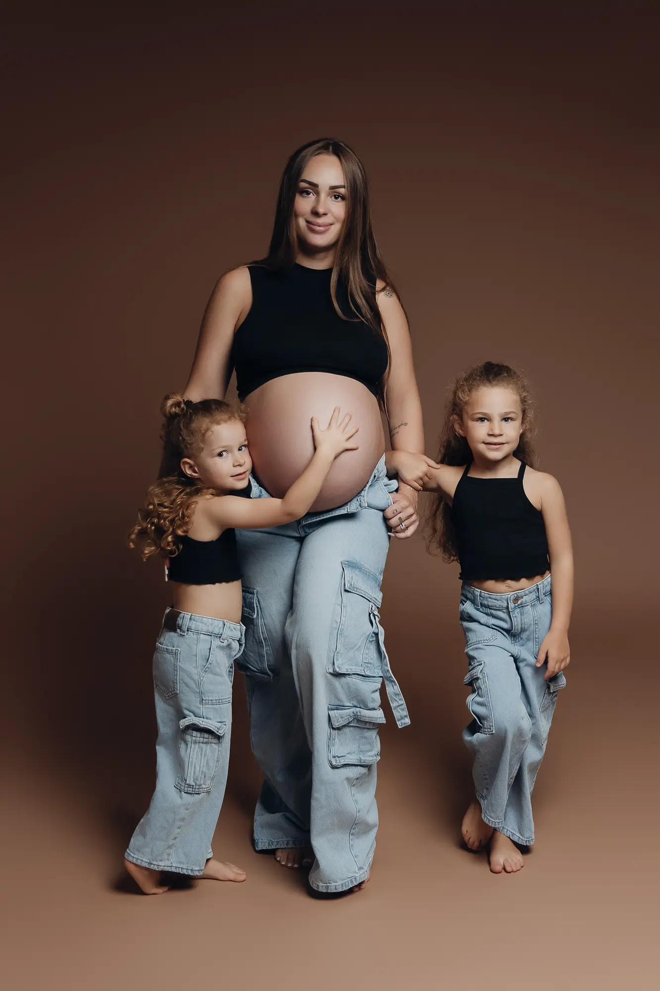 Grossesse maternelle au centre, entourée de deux jeunes filles en jeans, portrait studio sur fond brun. alt: Fille enceinte entourée de deux petites filles, studio grossesse bébé.