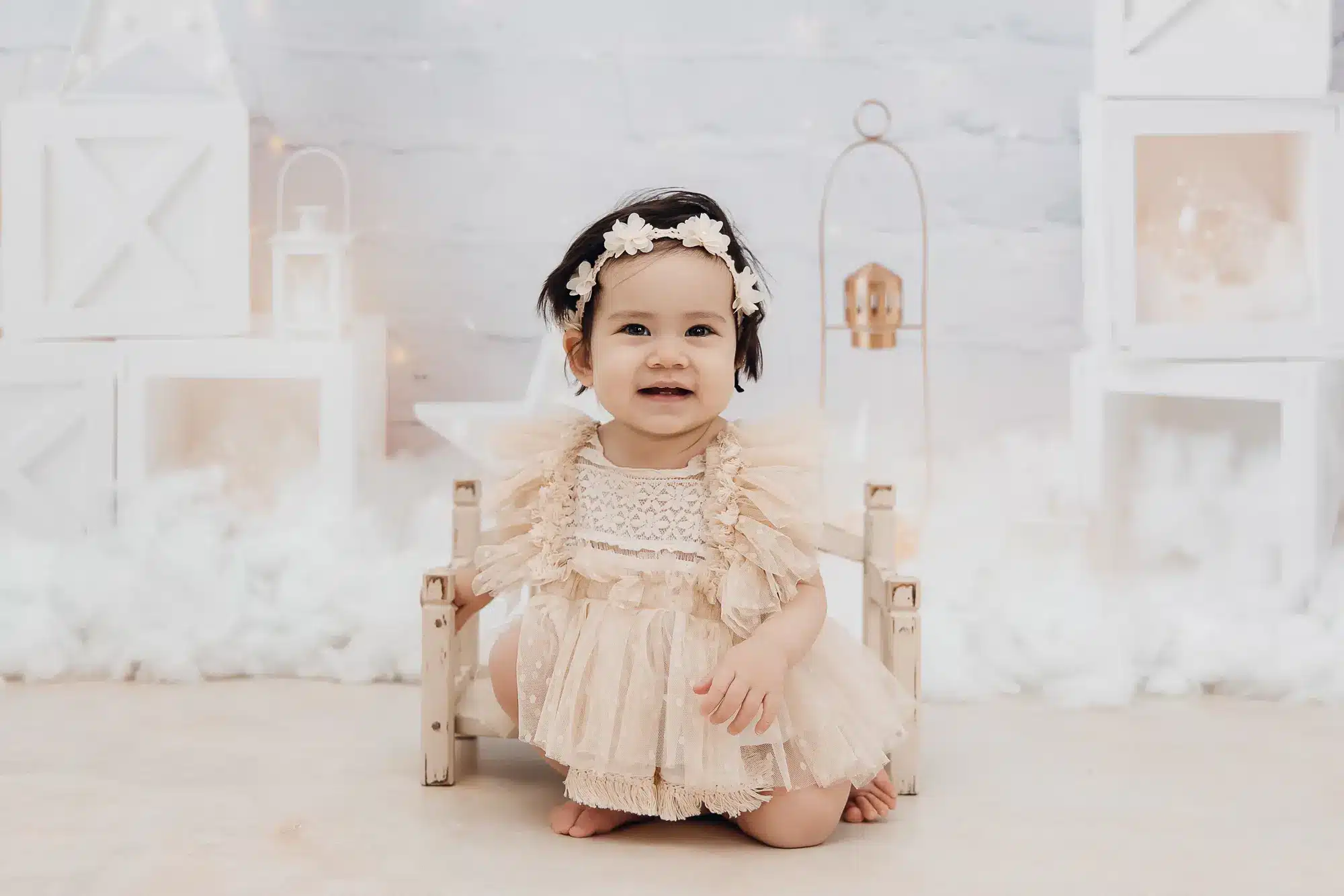 Petite fille assise sur un pouf, portant une robe délicate et une couronne de fleurs, décor pastel autour.