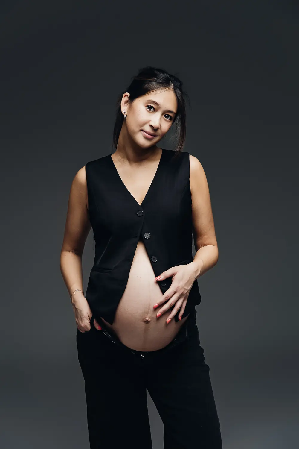 Photographie grossesse : femme enceinte debout, veston noir ouvert sur le ventre exposé, sourire léger, fond gris sombre.
