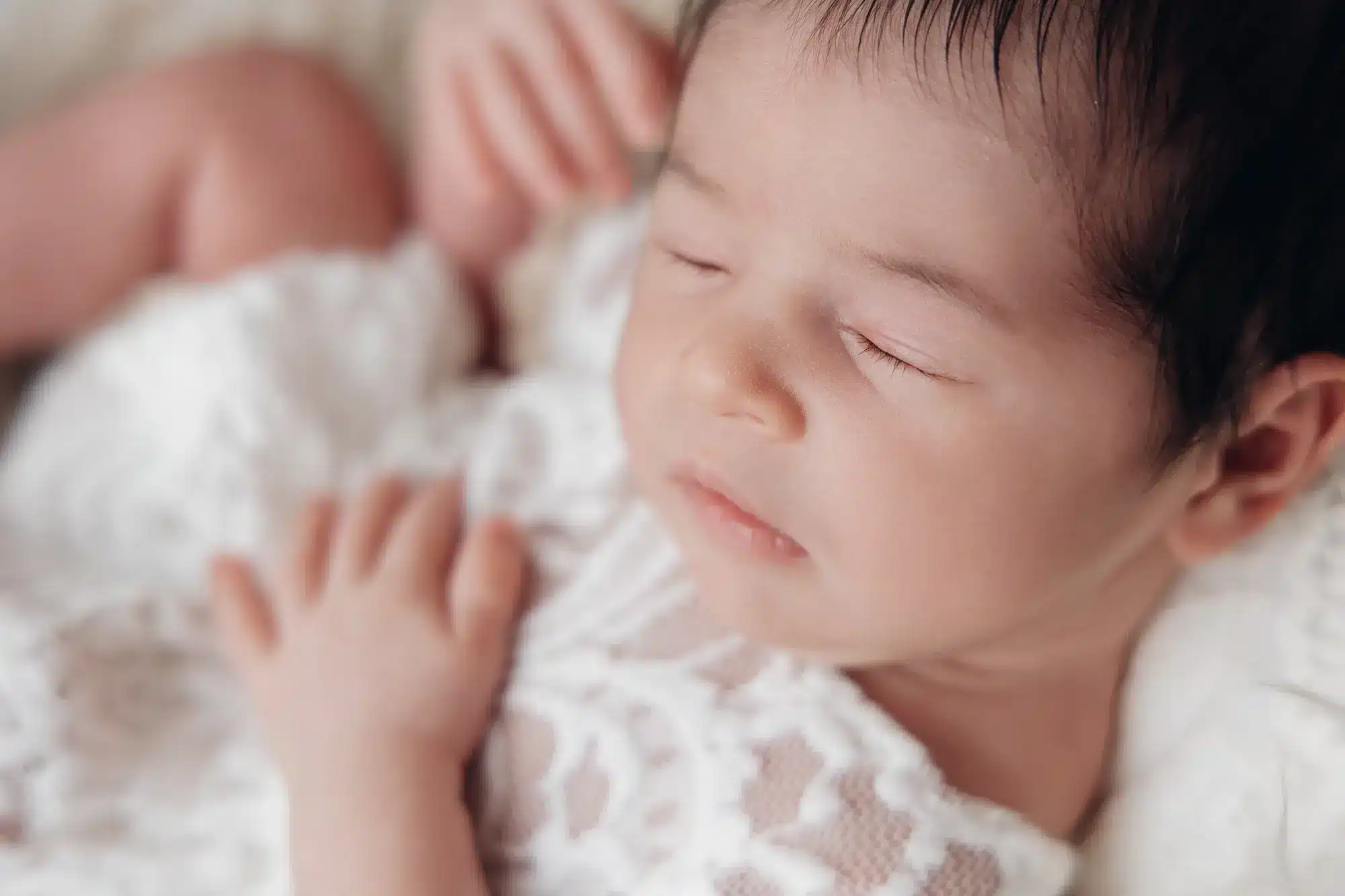 Bébé nouveau-né dormeur au visage calme, posé sur drap blanc délicat avec texture dentelle.