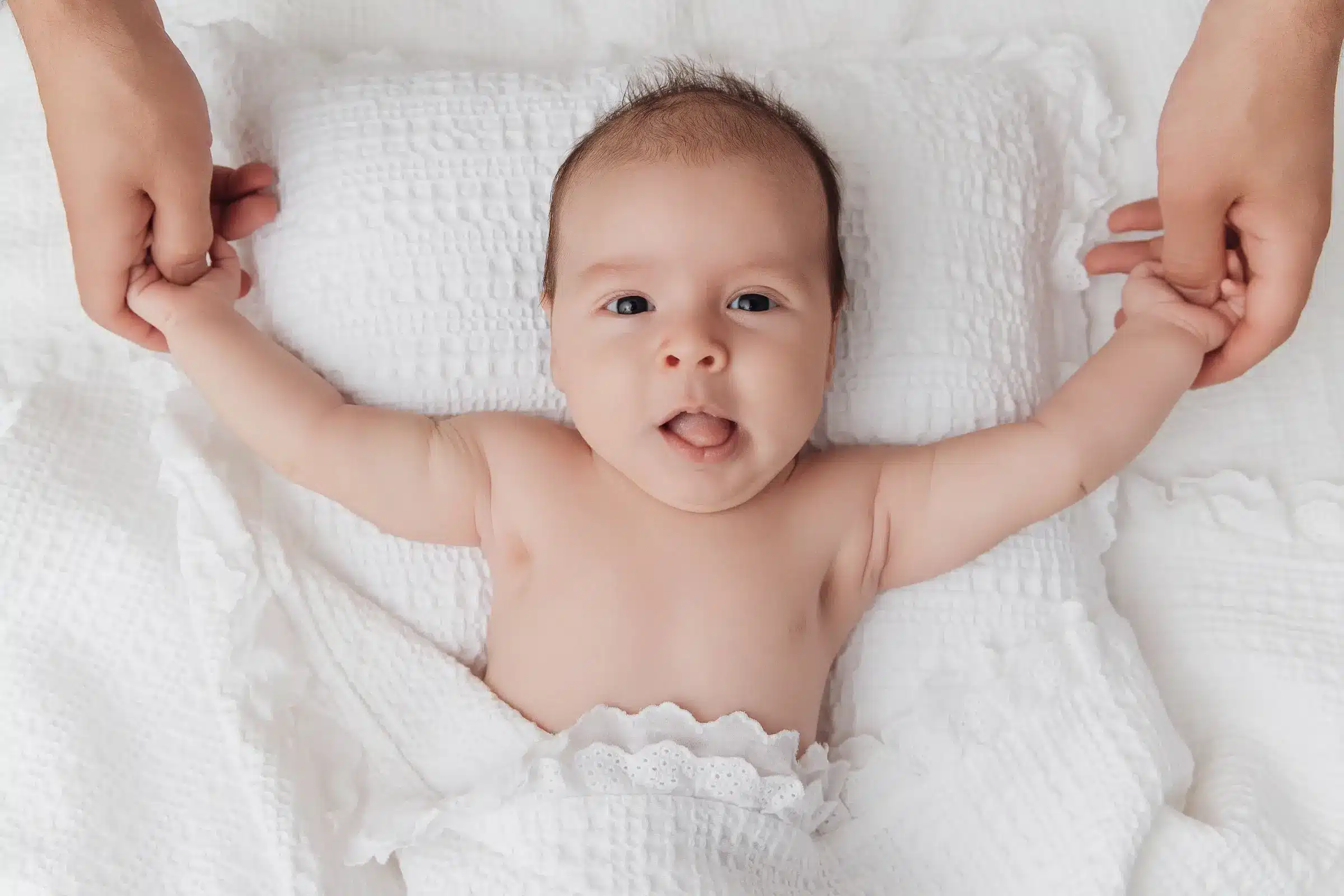 Bébé nu sur une couverture blanche, les bras tenus par deux mains adultes, visage souriant et langue légèrement sortie.