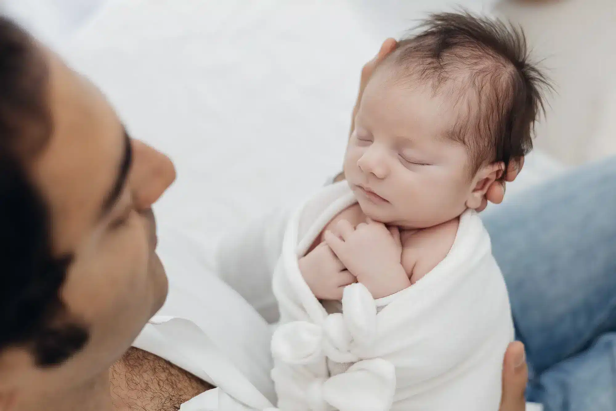 Belle premère prise en studio montrant un nouveau-né enveloppé, serein et dormant dans les bras d’un parent, avec une attention à l’hygiène et à la sécurité. hygiène en séance nouveau-né au cœur du regard.