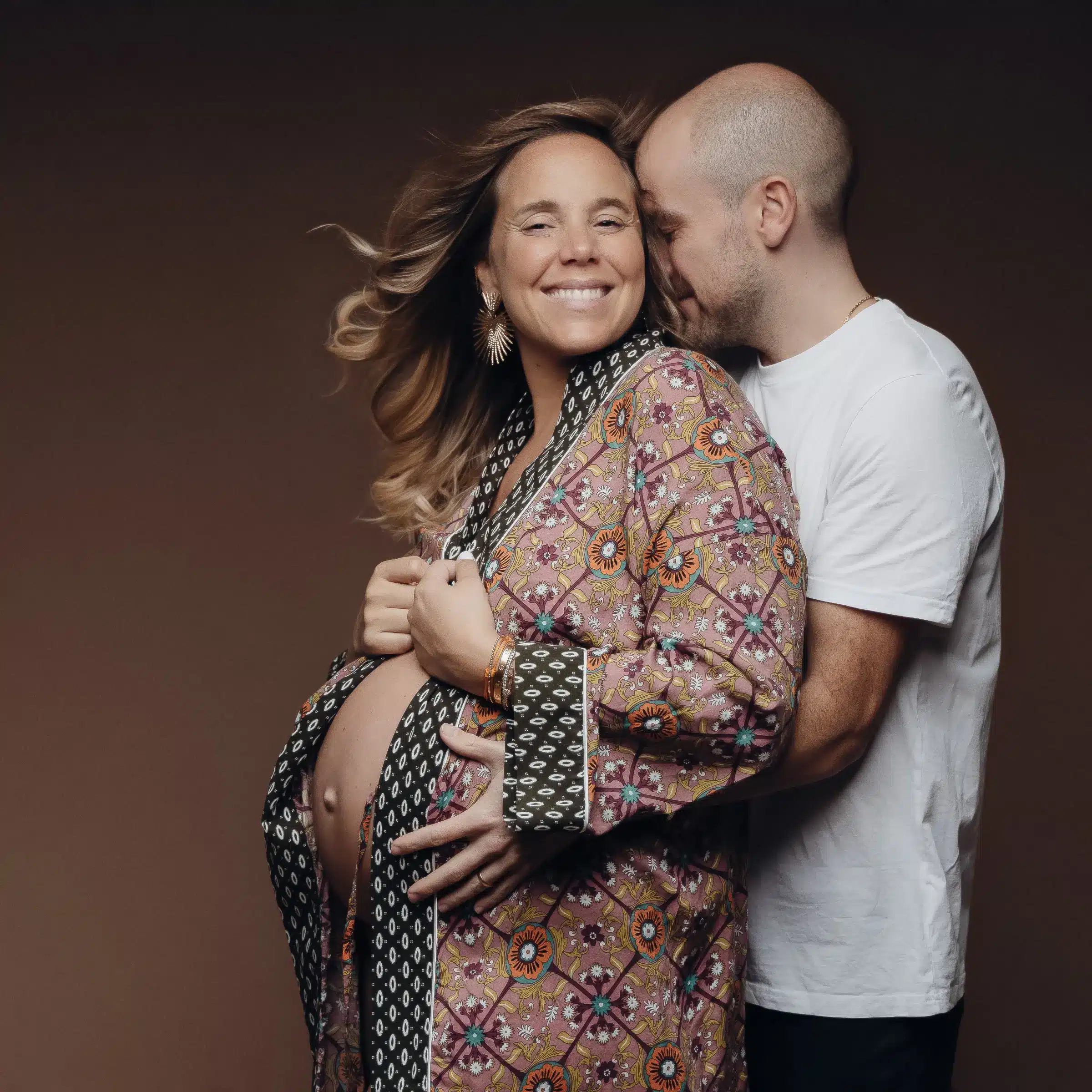 Couple en studio: femme enceinte souriante porte un kimono floral, homme derrière elle avec bras autour, gestes tendres sur ventre.