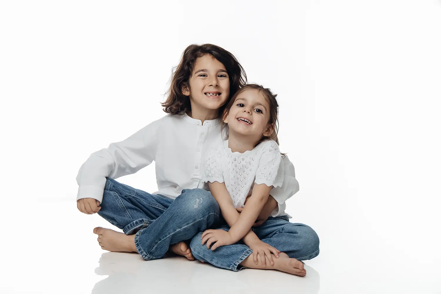 Deux jeunes frère et sœur assis par terre, souriants, jeans et chemises blanches sur fond clair.