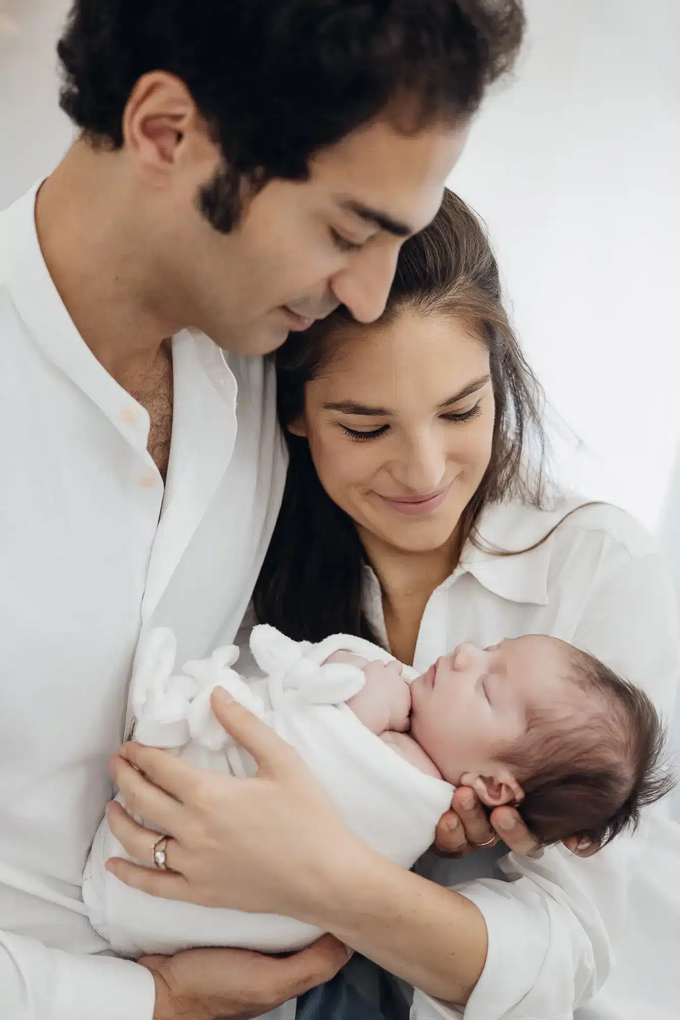 Famille joyeuse: un couple tient un nouveau-né enveloppé dans une couverture blanche, la mère et le père souriants et regardant tendrement le bébé. ALT: Famille et bébé newborn.