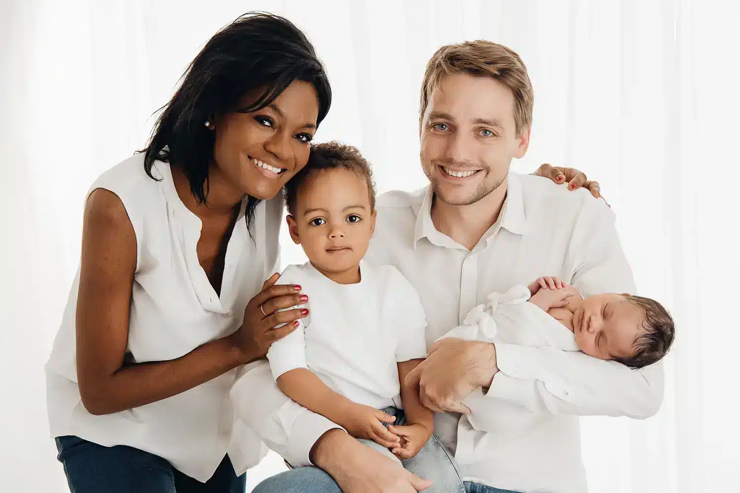 Une famille pose ensemble: deux parents souriants entourent deux jeunes enfants, l’aîné assis au centre et le bébé endormi dans les bras du père, dans une lumière douce. Comment réussir une photo de famille avec de jeunes enfants ?