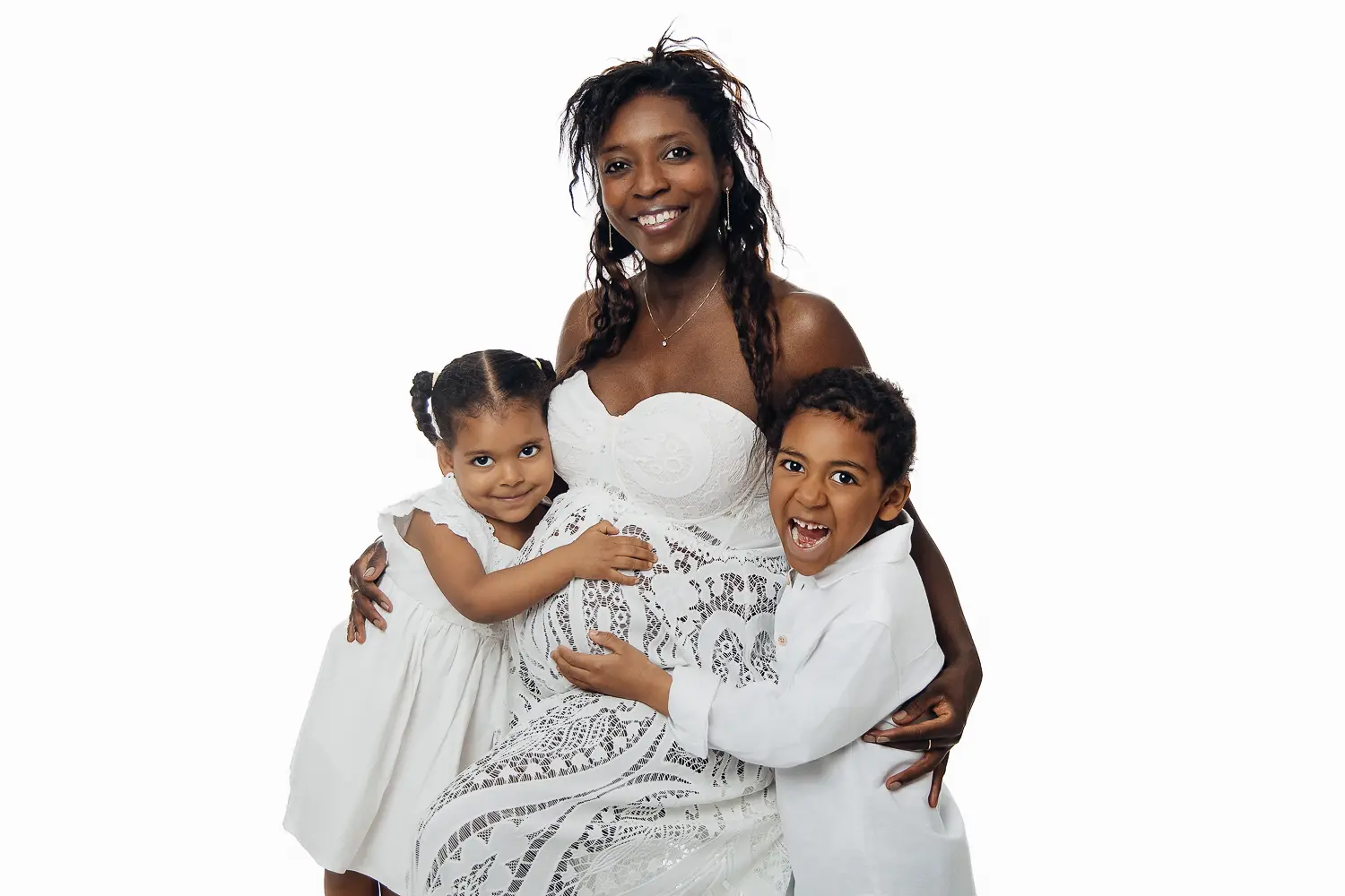 Famille rieuse et tendre: une mère noire entourée de deux enfants en robes blanches, tous souriants devant fond blanc. Faut-il forcer les enfants à sourire sur les photos ?