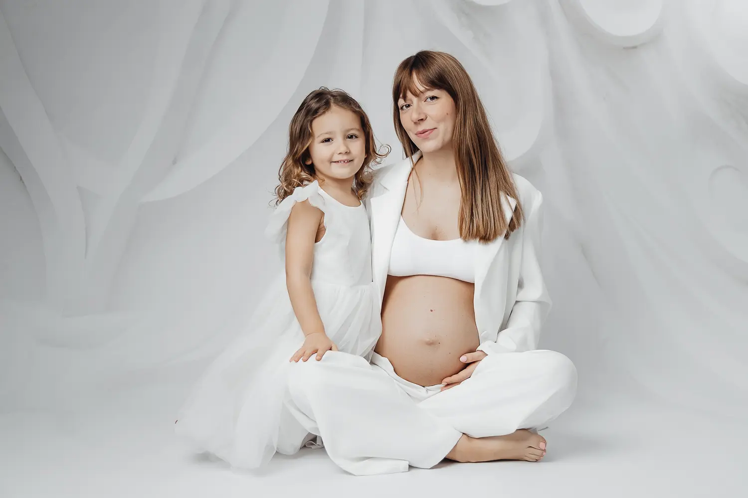 Une femme enceinte assise, les mains sur son ventre, accompagné d’une jeune fille en robe blanche, dans une ambiance studio blanche et grise. Une femme enceinte assise, les mains sur son ventre, accompagné d’une jeune fille en robe blanche, dans une ambiance studio blanche et grise.