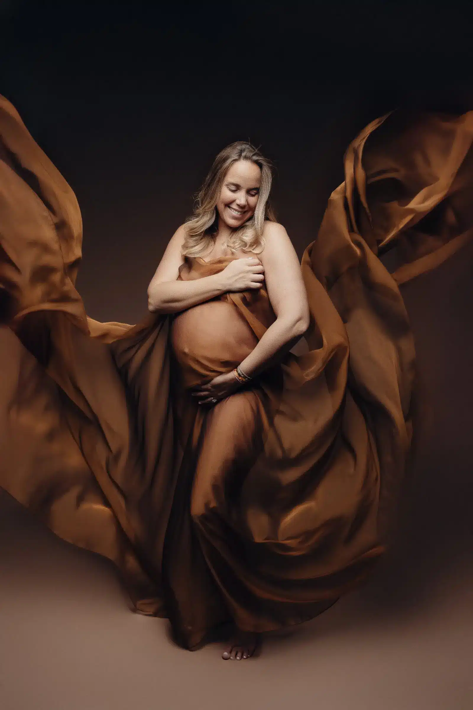Femme enceinte enveloppée d’un grand voile brun doré qui tourbillonne autour d’elle, posé en studio avec une atmosphère chaleureuse et lumineuse.