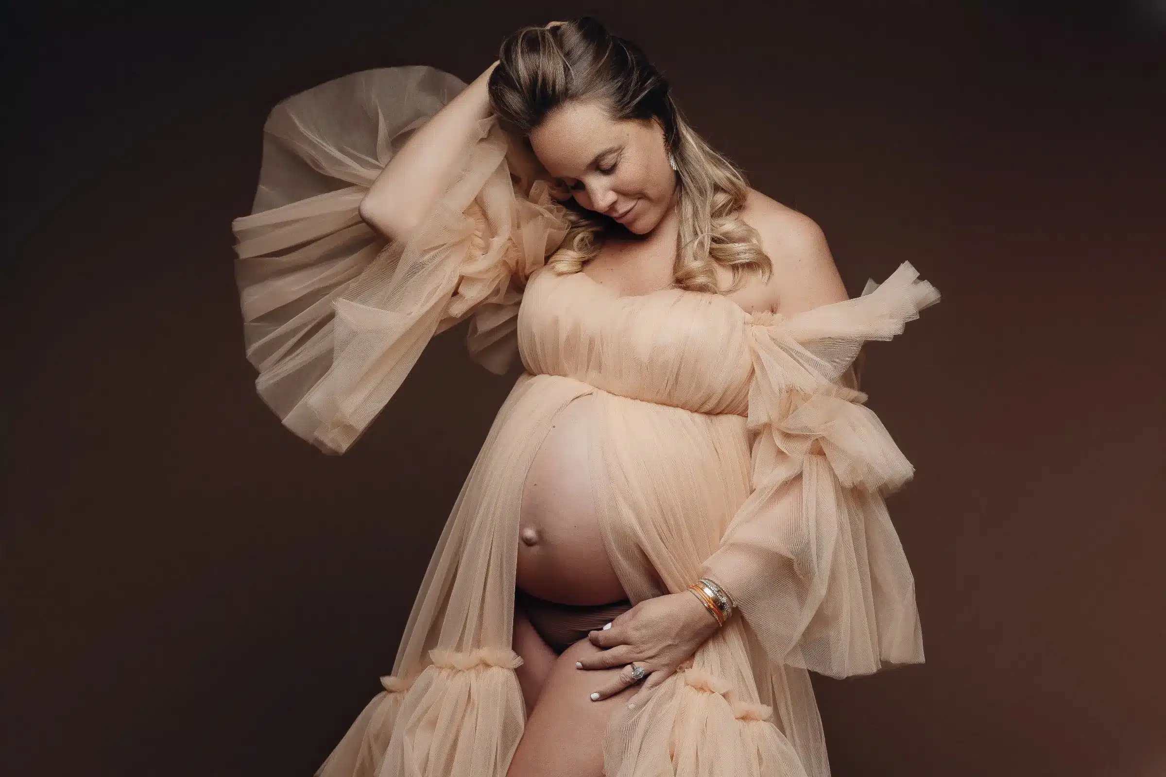 Femme enceinte posant en robe légère translucide, main sur le ventre et fond sombre, portrait maternité glamour.