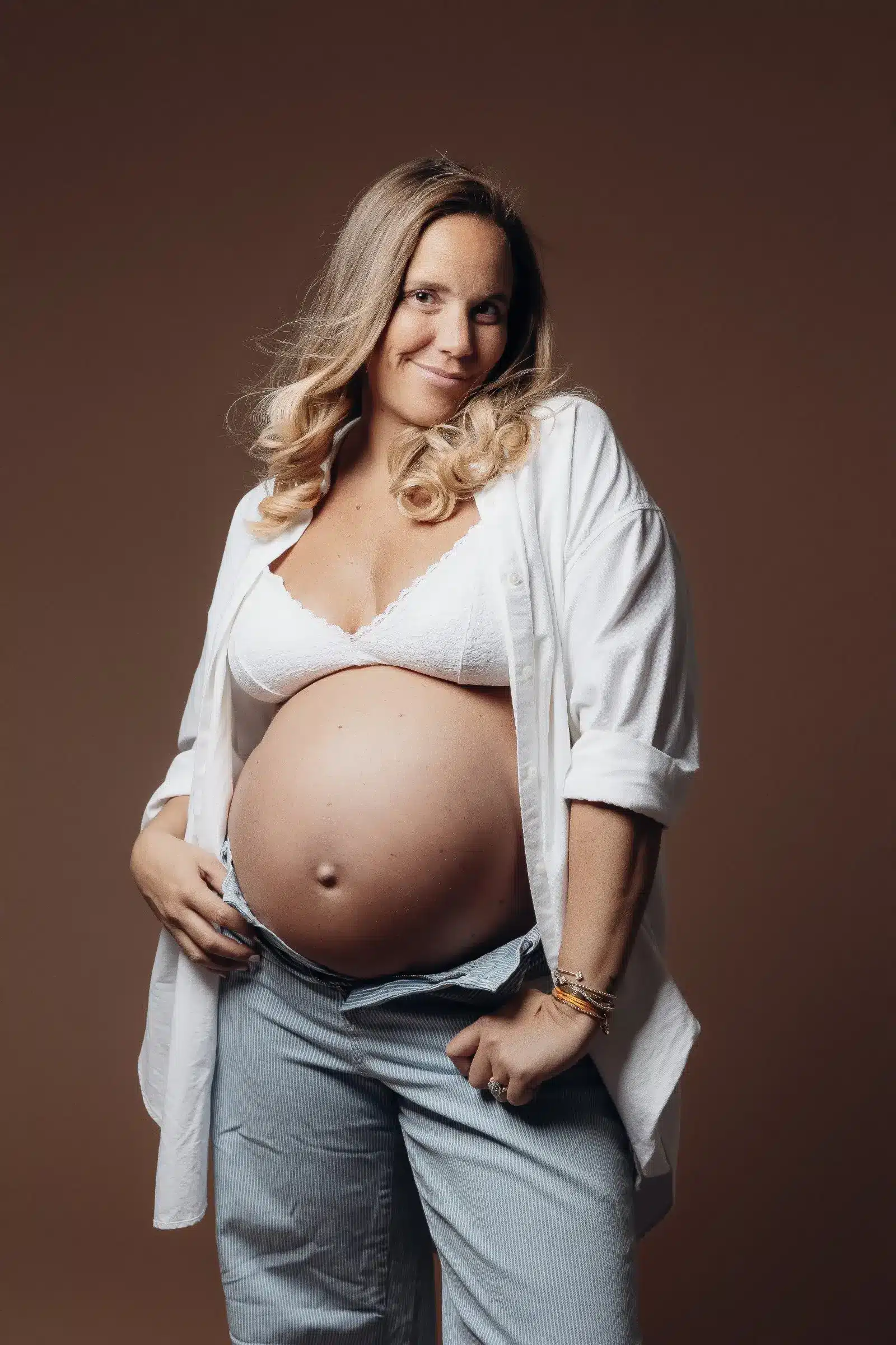 Femme enceinte rayonnante, portant chemise ouverte sur le ventre rond, lingerie blanche et pantalon léger, pose douce en studio. ALT: Femme enceinte pose dans un studio avec ventre proéminent.