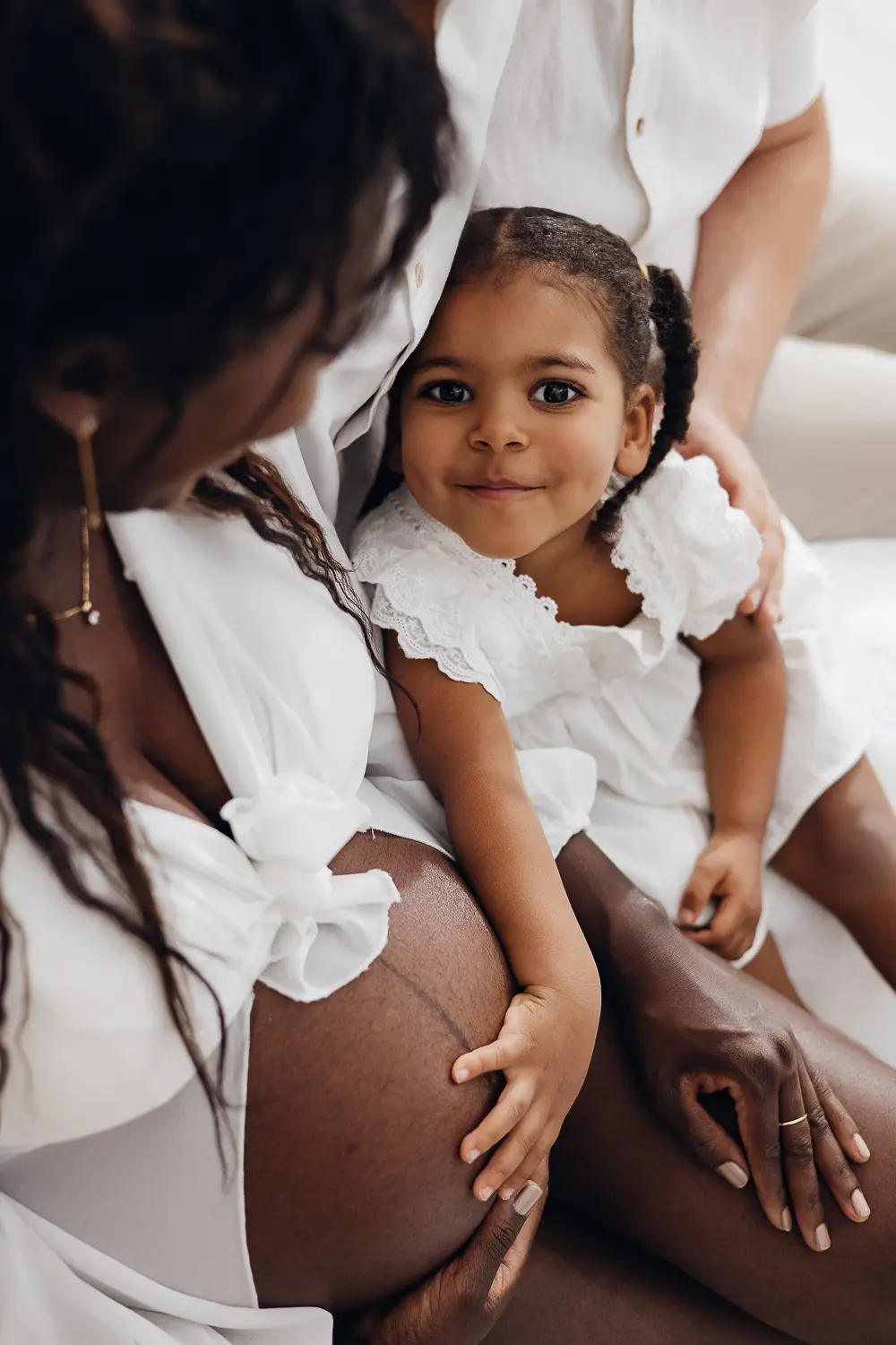 Gros plan sur une jeune femme enceinte caressant son ventre rond, avec une petite fille sur ses genoux et des mains qui soutiennent le ventre, ambiance tendrement familiale. POS.
