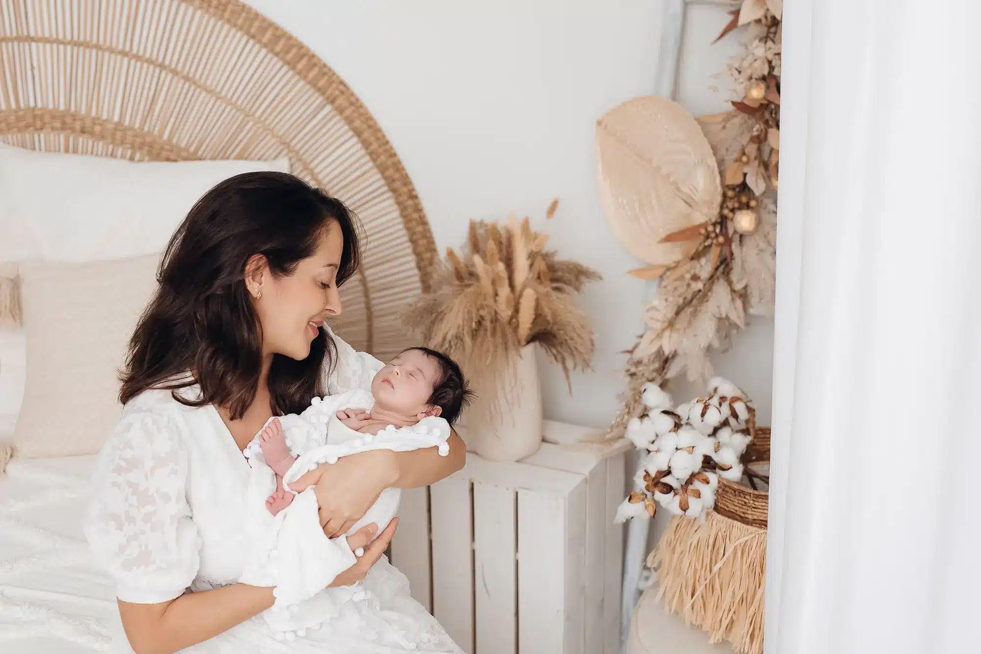 Méline, jeune mère, sourit en tenant son nouveau-né enveloppé dans une gigoteuse blanche dans un studio décoré de pampas et d’éléments naturels.