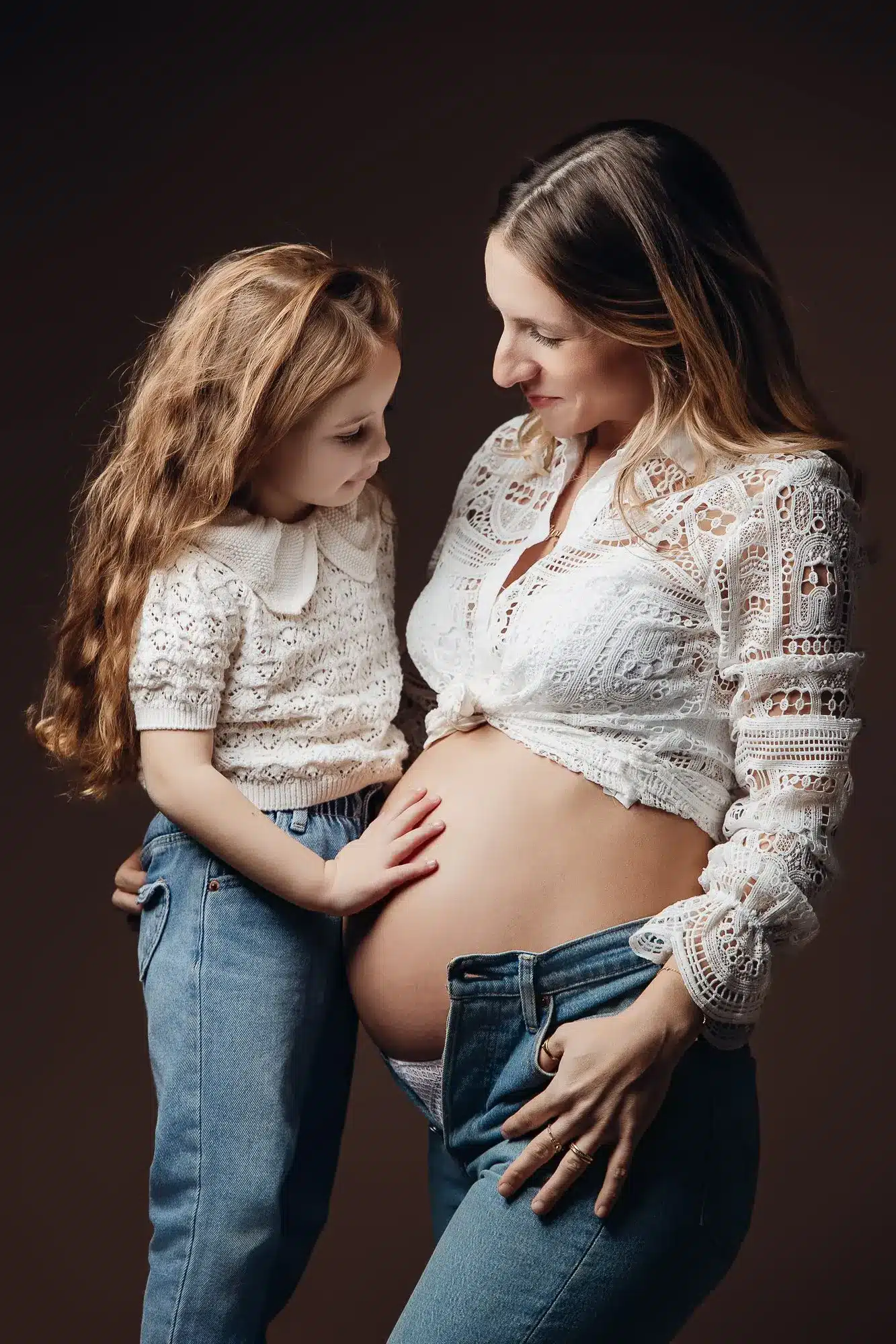 Mère enceinte et jeune fille se touchant le ventre, dans une séance grossesse intimiste dans des tenues blanches.