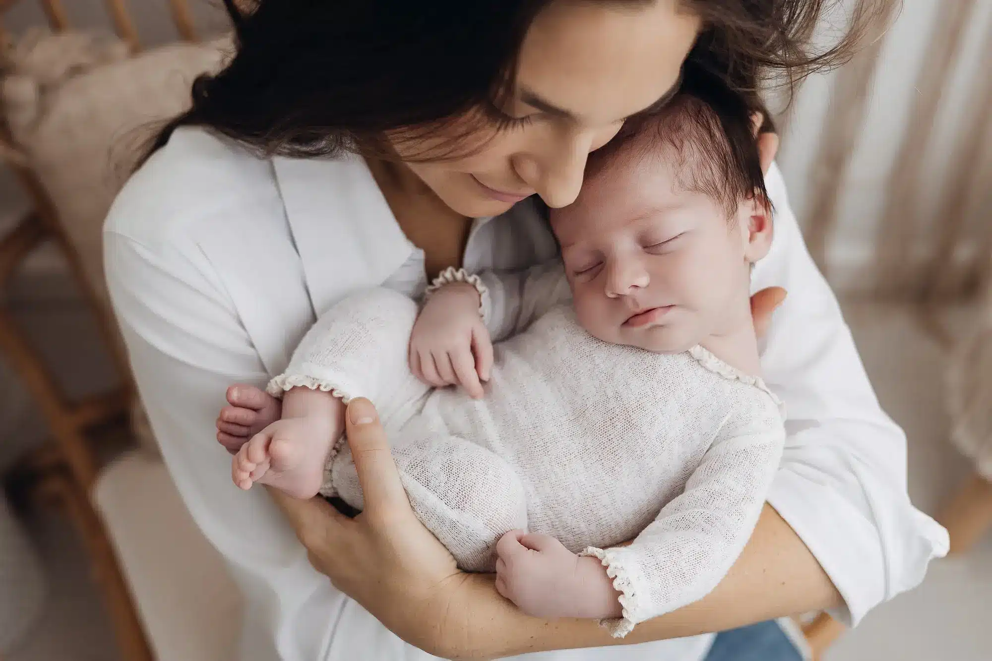 Mère souriante tient son nouveau-né endormi contre elle dans un cadre domestique, bébé enveloppé dans une tenue légère blanche.