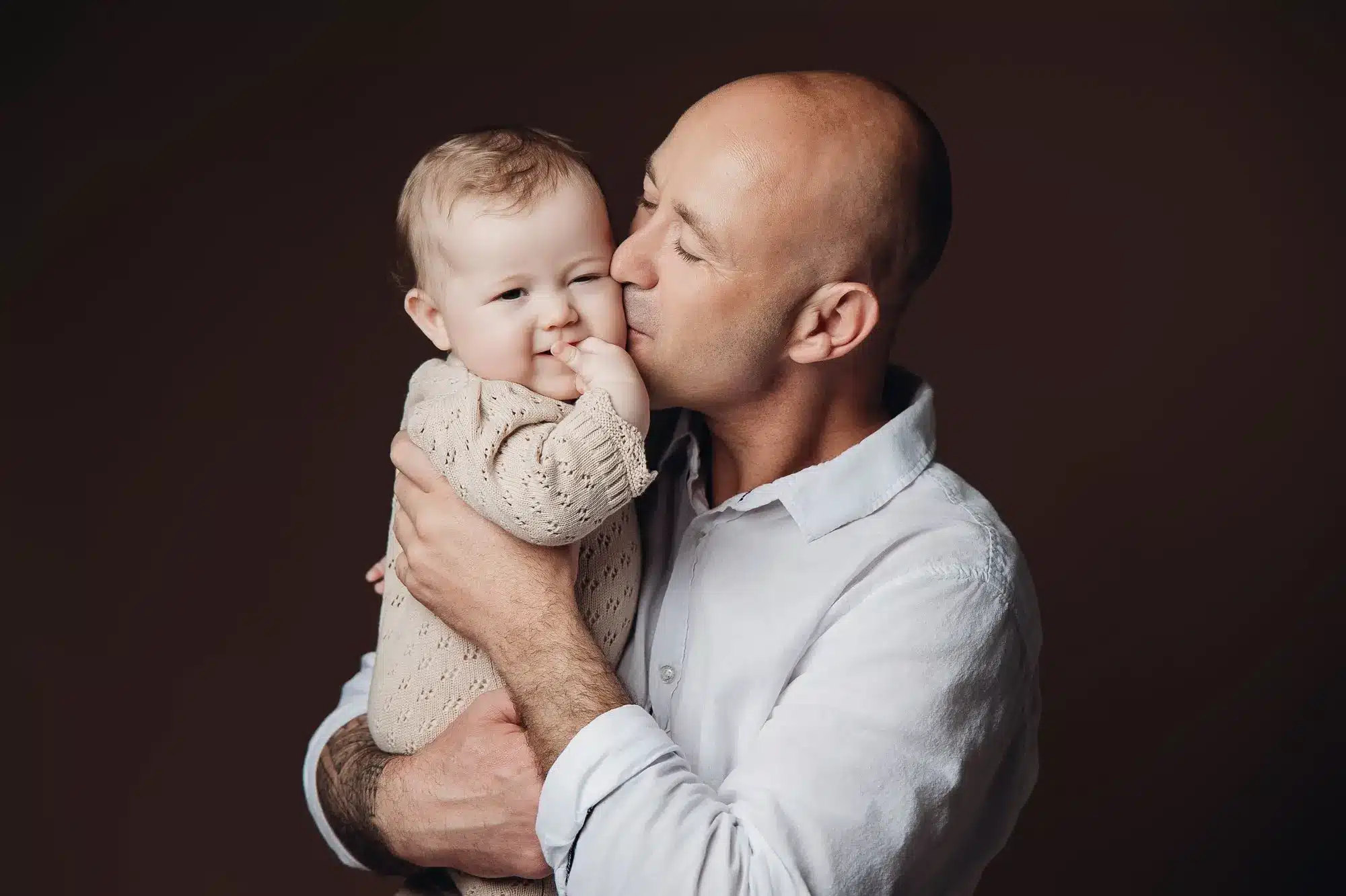 Père embrassant tendrement son bébé, dans une scène chaleureuse et intime, éclairage doux et fond sombre.