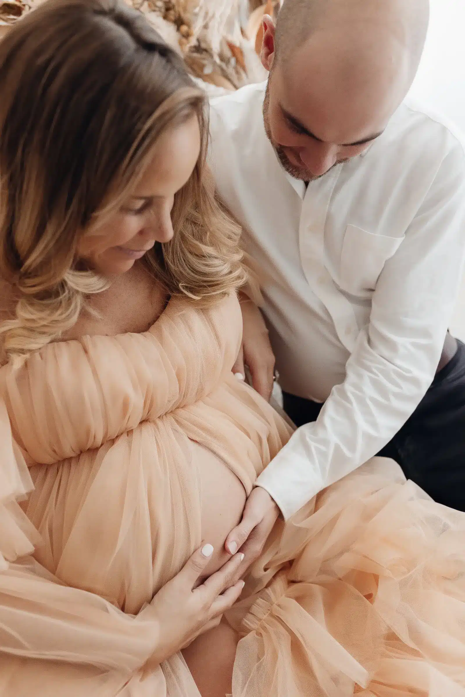 Photo de grossesse: une femme enceinte en robe beige flottante, son partenaire touche son ventre rond, dans une atmosphère tendre et intime.