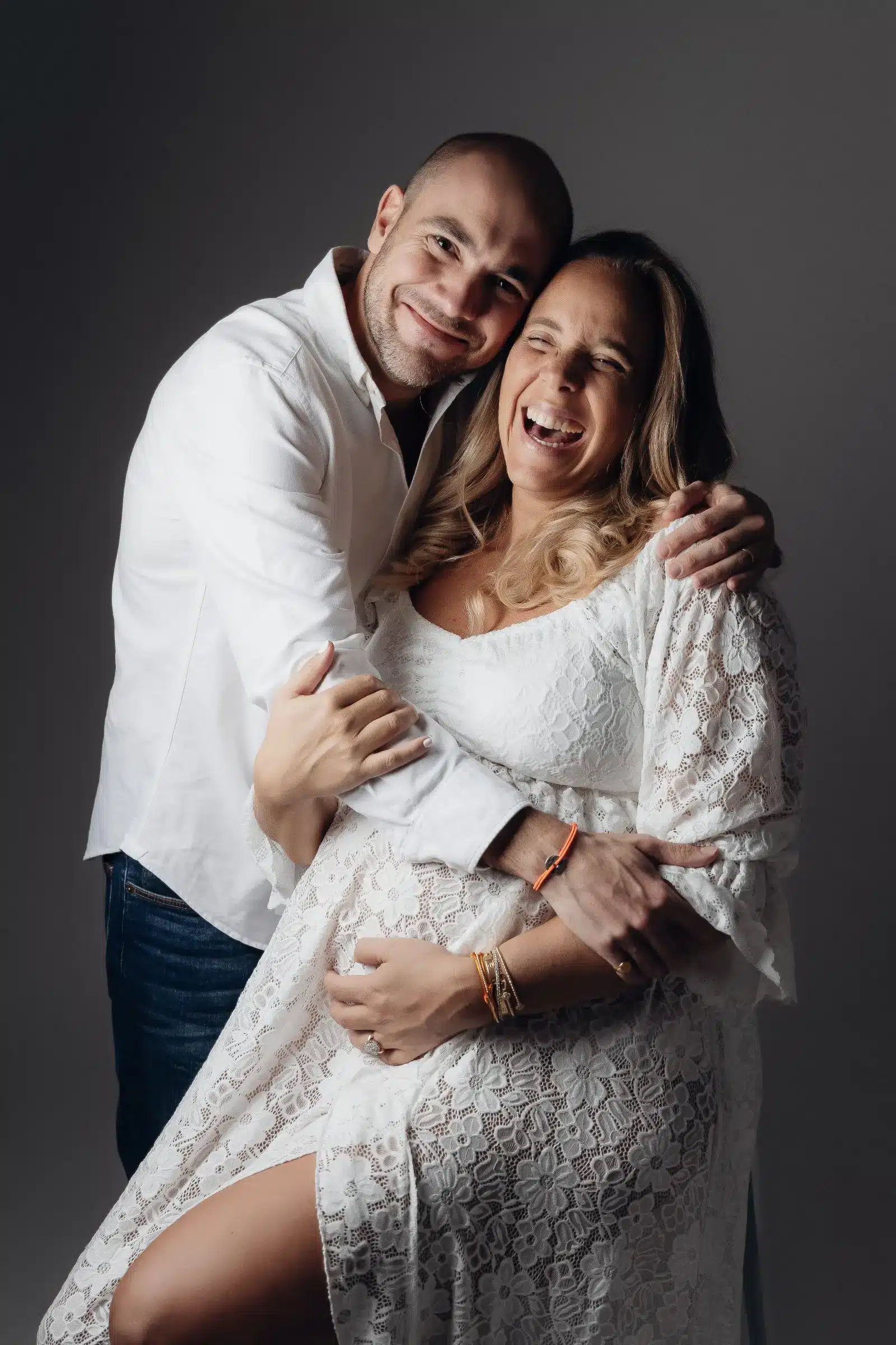 Portrait chaleureux d'un couple souriant, maman enceinte en robe lace et papa en chemise blanche, se serrant dans les bras.