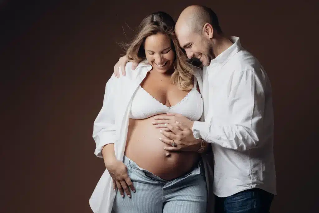 Portrait de couple avant l'arrivée de bébé: femme enceinte, ventre prononcé, mains sur le ventre et bébé dans les bras, ambiance chaleureuse.