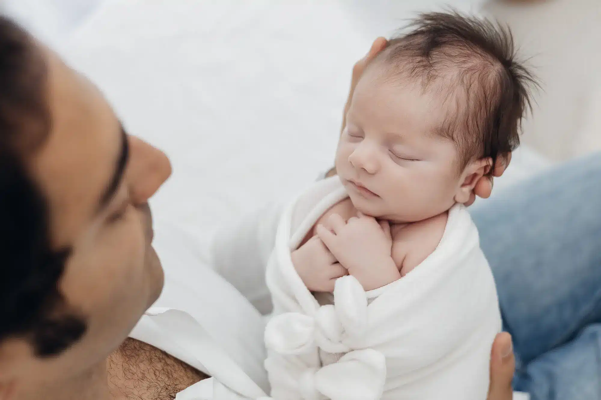Portrait d’un nouveau-né endormi, enveloppé dans une couverture blanche, tenu tendrement par un parent. Alt: bébé dormant emmaillotté.