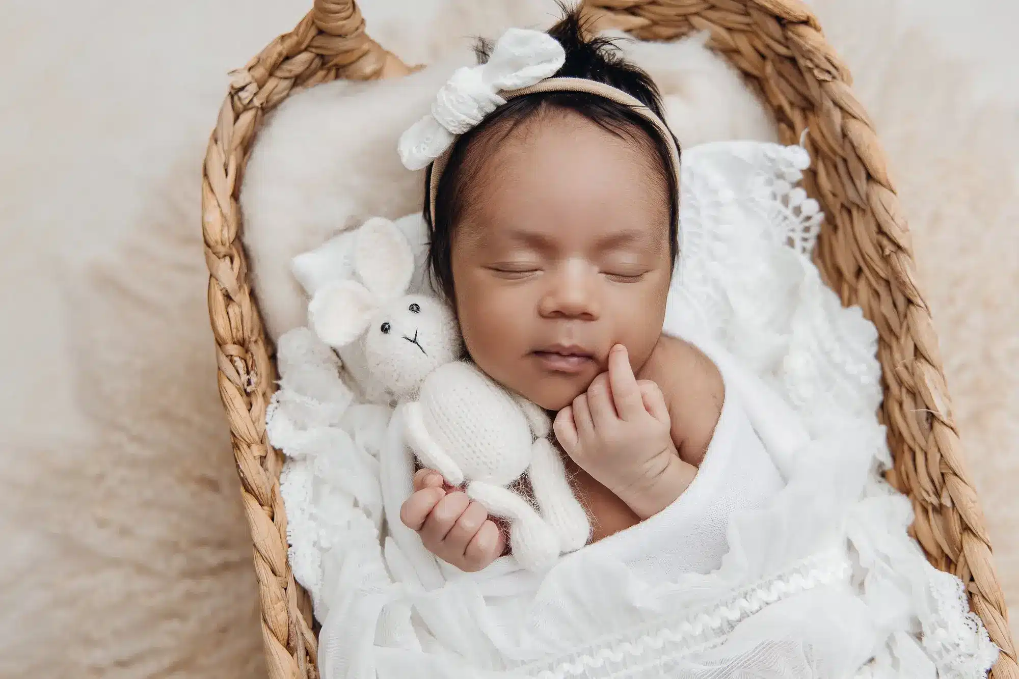 Nouveau-né endormi dans un berceau en osier, enveloppé de linge blanc avec un petit ours en peluche blanc et un bandeau blanc sur la tête.
