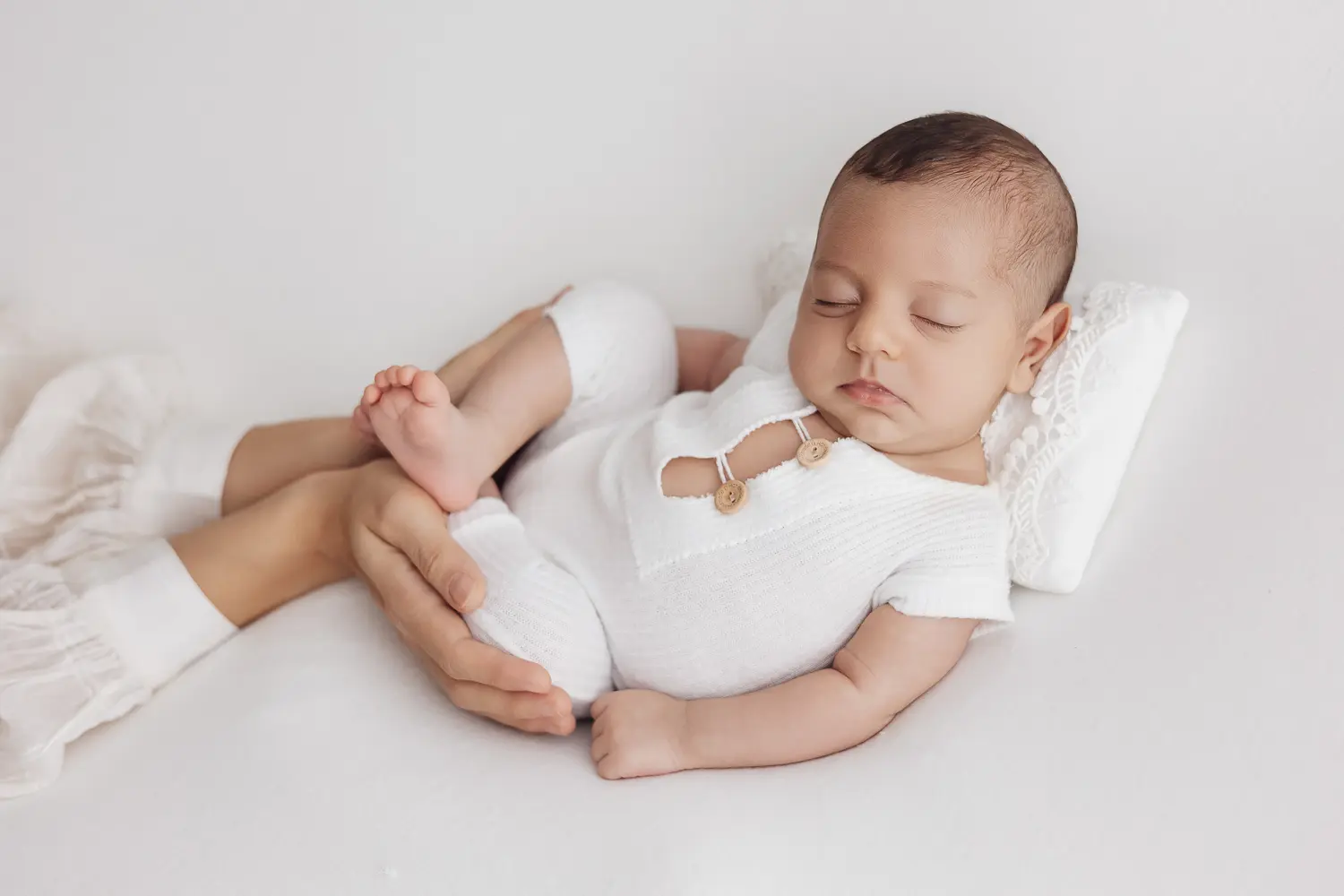 La sécurité en séance photo nouveau-né : bébé endormi sur le dos, entouré par des mains douces, vêtements blancs et peluche légère.